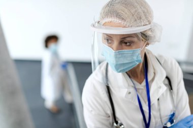 Koronavirüs salgını nedeniyle hastanede maske ve güneşlik takarken kolları çapraz duran dalgın bir sağlık görevlisi.. 