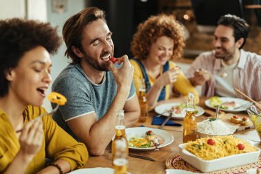 Happy man eating cherry tomato while having lunch with friends at dining table at home. 