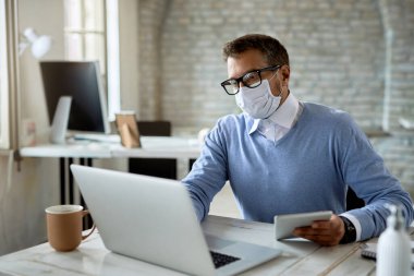 Male entrepreneur with protective face mask using touchpad while working on laptop in the office. 