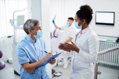 Dental assistant taking notes while communicating with black female dentist at dental clinic. 