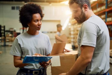 African American warehouse worker talking to her colleague while working at industrial storage compartment. 