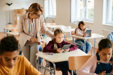 Mature teacher assisting her elementary student in using digital tablet during computer class at school.