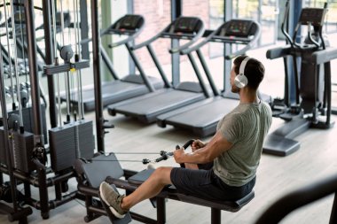 Athletic man with headphones exercising on rowing machine in a gym.