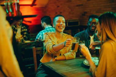 Group of young happy people toasting with beer and having fun at night in a pub. Focus is on Black woman. 