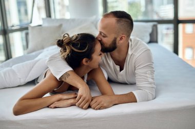 Young couple in love lying down in bed. Man is kissing his girlfriend on forehead. 