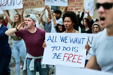 Large group of displeased protesters participating in public demonstrations against racism. Focus is on black woman holding a banner. 