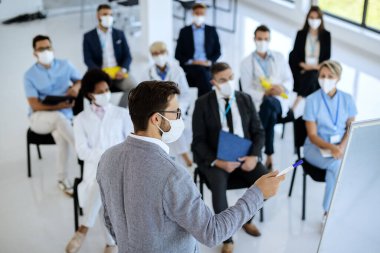 Businessman giving a presentation and wearing protective face mask while talking to group of entrepreneurs and healthcare workers in convention center.