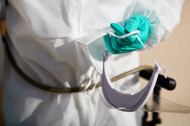 Close-up of healthcare worker wearing protective suit and gloves while holding visor and KN95 face mask.