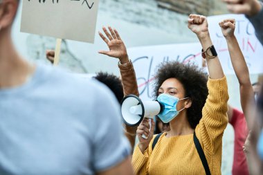 Large group of protesters with raised fists participating in demonstrations during COVID-19 epidemic. Focus is on black woman with protective face mask shouting through megaphone. 