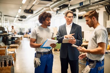 Mid adult engineer using touchpad while talking with steel workers in industrial building. 