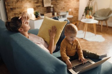 Stressed mother holding her head in pain while her small son is using computer at home.