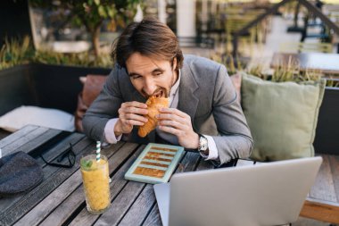 Hungry businessman reading an e0mail on laptop while eating croissant on lunch break in a cafe. 