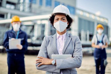 African American building contractor with protective face mask standing with her arms crossed and looking at the camera. Two civil engineers are in the background. 