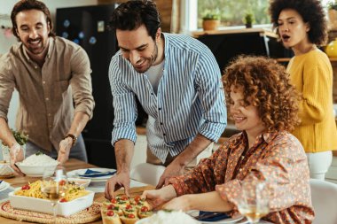 Young happy man and his friends bringing food at dining table while having lunch together at home. 