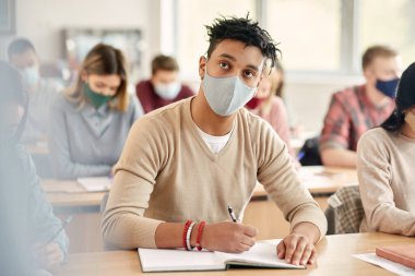 Young African American student writing in notebook while attending university classes during coronavirus pandemic.
