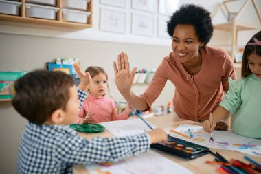 Happy African American teacher and small boy giving high-five during art class at kindergarten. 