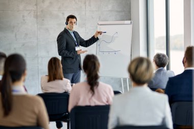 Male CEO wearing protective face mask while giving presentation to group of business people in board room.