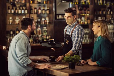 Happy waiter talking to a couple and serving them coffee while wearing protective face mask in cafe.