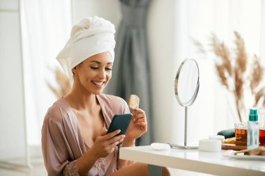 Genç mutlu kadın akıllı telefon kullanıyor ve evde cildine baktıktan sonra bir şeyler atıştırıyor..