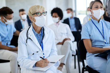 Mature female doctor wearing protective face mask and writing notes while attending educational event at conference hall. 