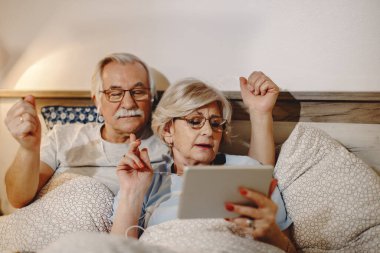 Senior woman singing while listening music with her husband over digital tablet in bedroom. 