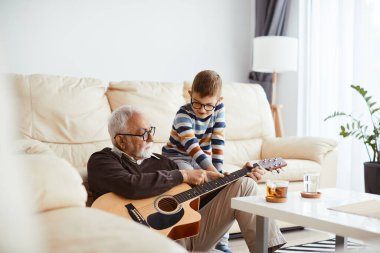 Senior man enjoying with his grandson and teaching him to play acoustic guitar at home. 