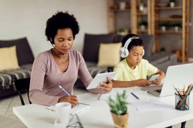 African American mother using digital tablet while writing notes and working at home. Her son is using mobile phone while sitting next to her. 