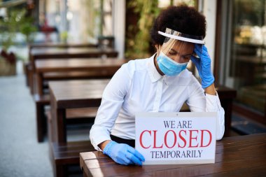 Black waitress holding closed sign and feeling worried about going out of business due to coronavirus pandemic. 