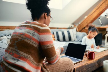 Evrakları analiz eden ve kızıyla evdeyken dizüstü bilgisayar kullanan Afrikalı Amerikalı bir annenin arka planı.. 