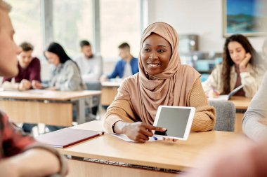 Happy African American Islamic student and her friend e-learning on touchpad during a lecture at the university.