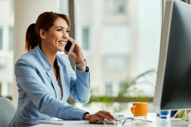 Young happy businesswoman working on desktop PC and communicating over mobile phone in the office.