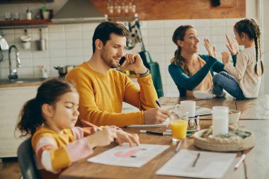 Smiling parents with two daughters at home. Focus is on working father talking on the phone and takin notes. 