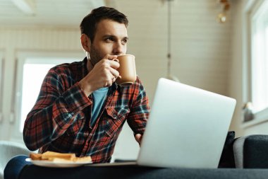 Low angle view of young man thinking while drinking coffee and using computer in the living room. 