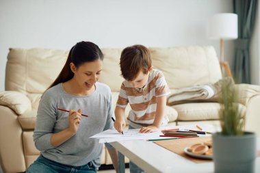 Young mother having fun and drawing on the paper with her son at home.