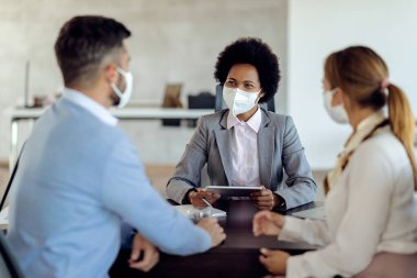 Happy African American financial consultant and her clients wearing protective face masks while having a meeting in the office. 