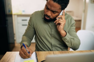 Black man taking notes while communicating over mobile phone and working at home. 
