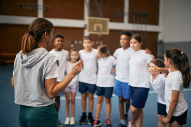 Back view of female PE teacher explaining exercise activities to group of kids at school gym 