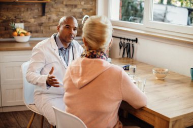 Black general practitioner talking to mature woman while visiting her at home. 