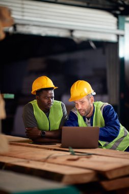 Manual workers talking while working on a computer at lumber warehouse.