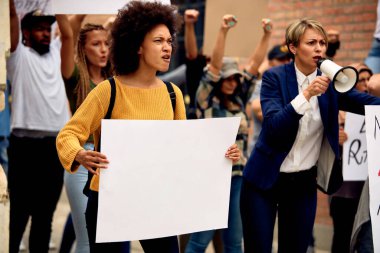 Large group of displeased protesters marching at demonstrations on the streets. Focus is on African American woman holding blank placard. Copy space.