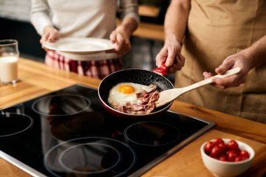 Close-up of couple having breakfast at home. Man is preparing eggs and bacon in frying pan. 