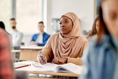 African American Islamic student taking notes while attending a lecture at college classroom.