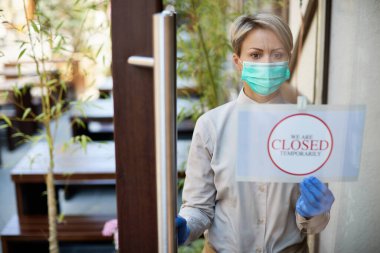 Female cafe owner hanging closed sign on entrance door due to COVID-19 pandemic. The view is through the glass. 