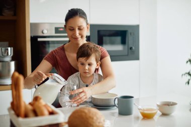 Young mother pouring milk into a glass while having breakfast with her son at home. 