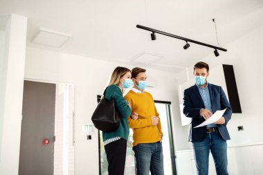 Low angle view of young couple and their real estate agent examining housing plans while wearing face masks due to coronavirus pandemic. 