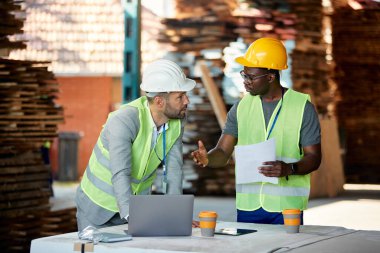 Mid adult businessman using laptop and talking to African American worker while analyzing paperwork at lumber warehouse. 