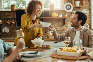 Young happy woman talking to her friend while bringing food for lunch at dining table. 