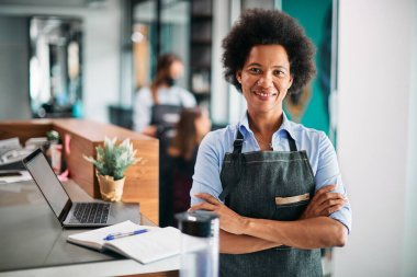 Confident black hairdresser standing at reception desk at her salon and looking at camera. 