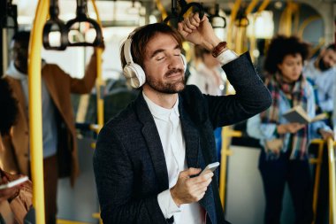 Smiling entrepreneur using smart phone and listening music over headphones while traveling to work by bus. 