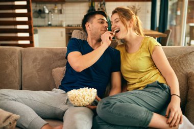 Happy woman having fun while boyfriend is feeding her with popcorn in the living room. 